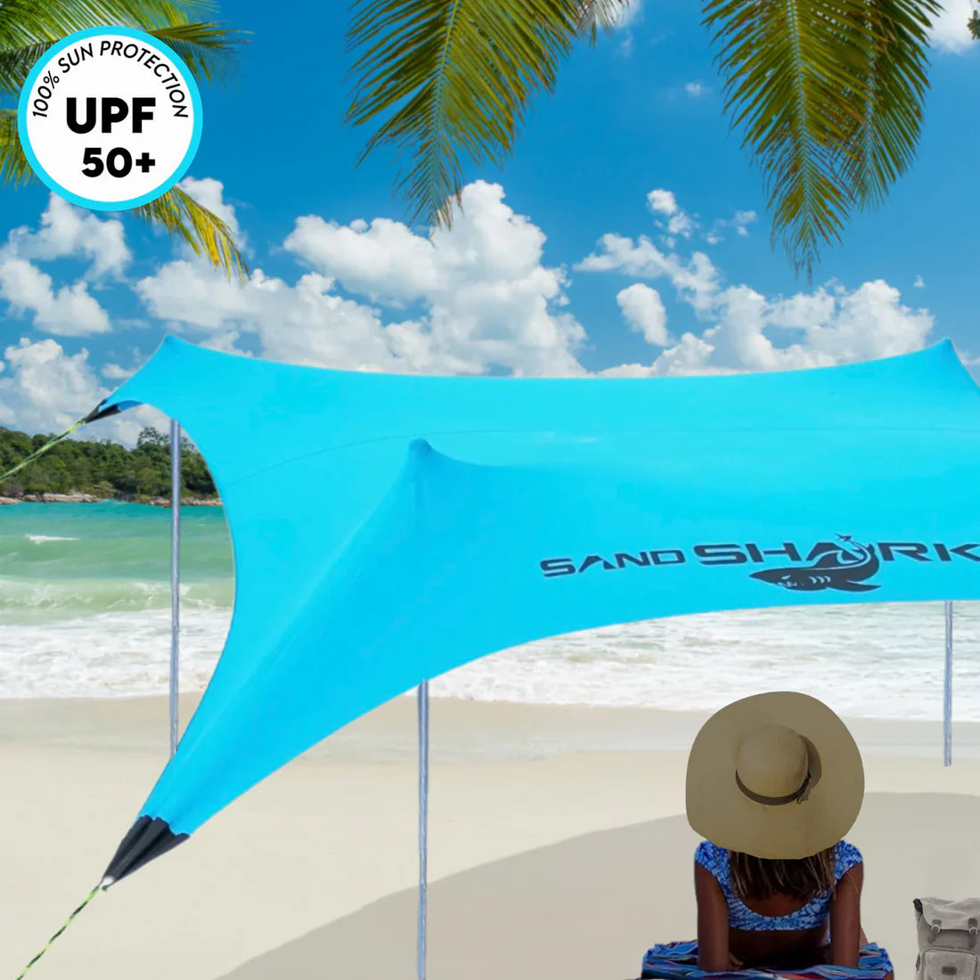 Blue SandShark sun shelter on a beach with a person sitting underneath, palm trees, and ocean in the background.