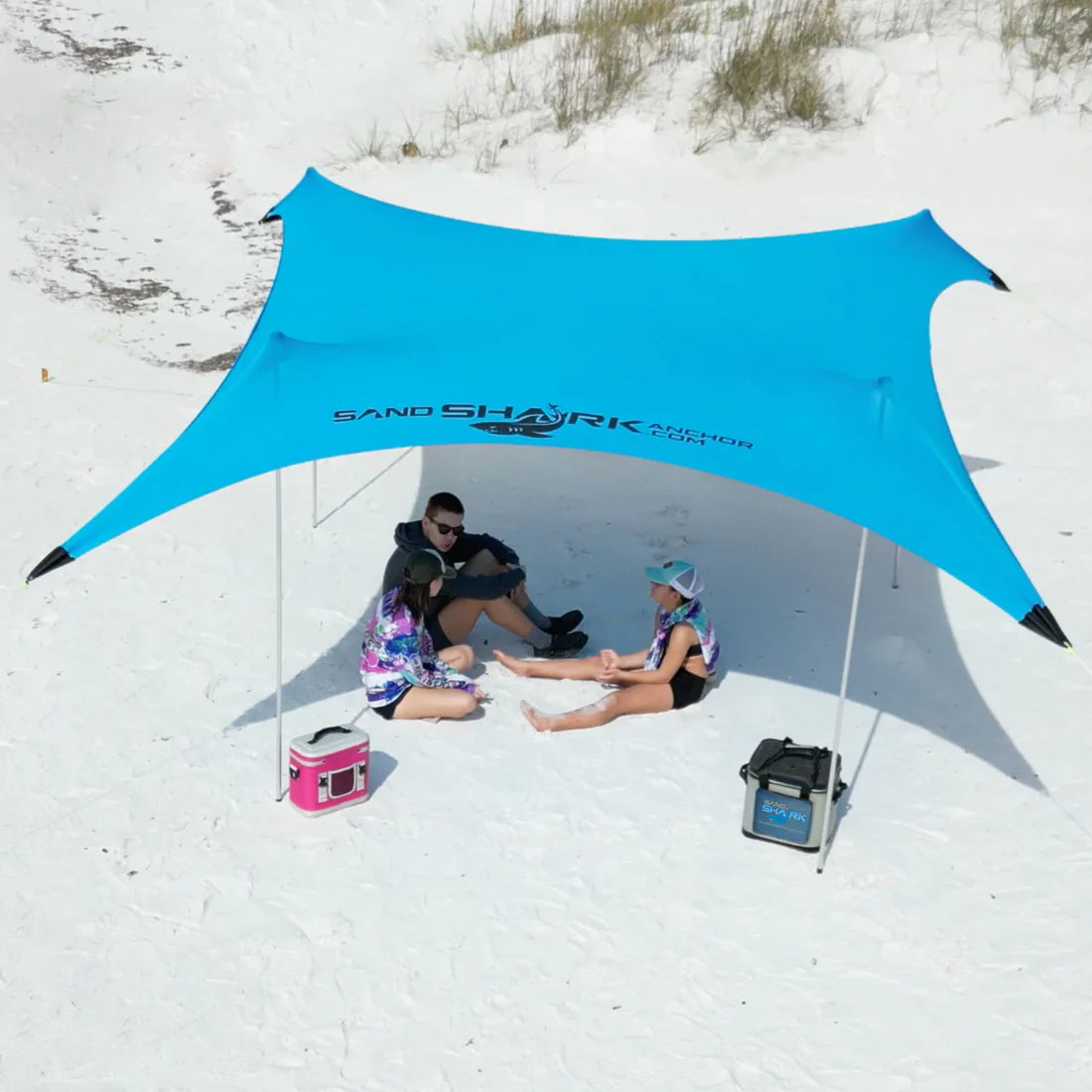 People sitting under a blue SandShark canopy on a sandy beach.