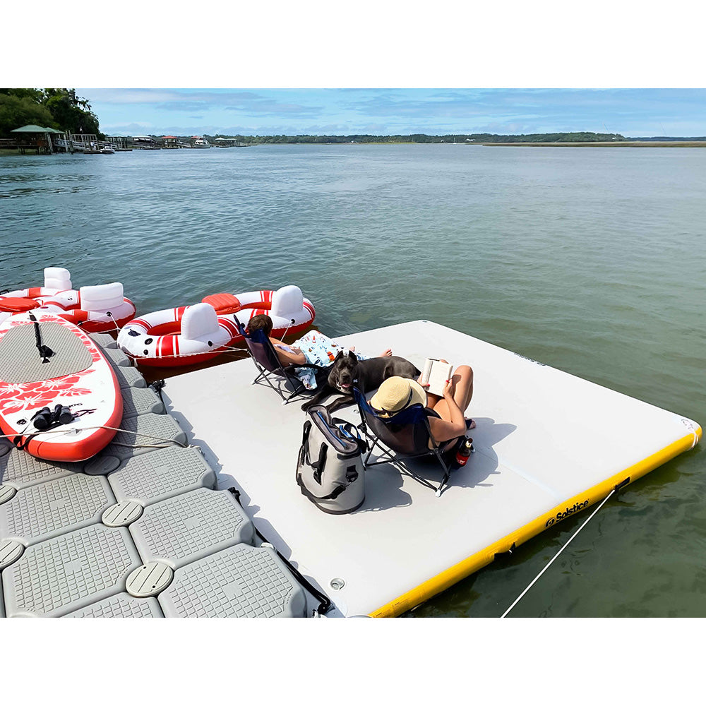 Two people lounging on a Solstice inflatable dock platform with chairs, cooler, and paddleboard on a calm lake