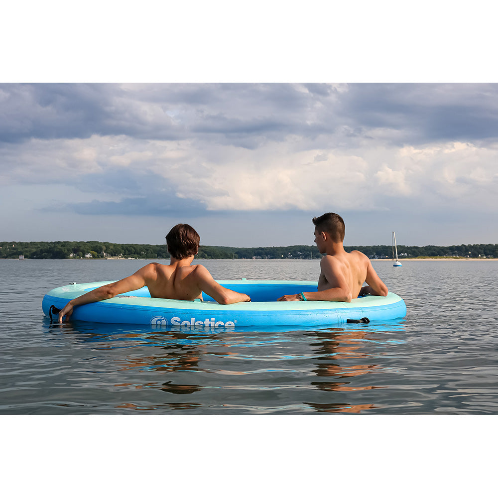 Two people lounging in a blue inflatable floating ring on calm lake water with cloudy sky in the background”