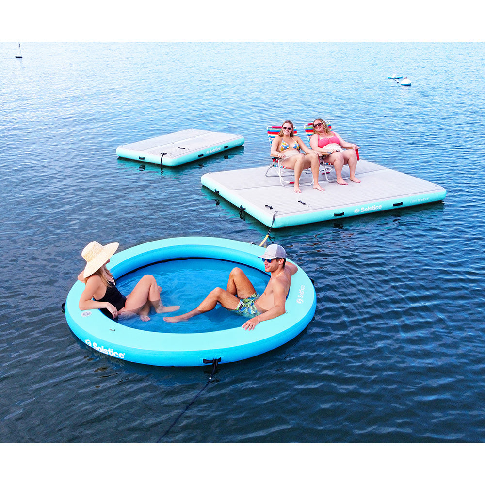 “Two people relaxing in a blue inflatable water lounge near two Solstice floating docks with people sitting in chairs”